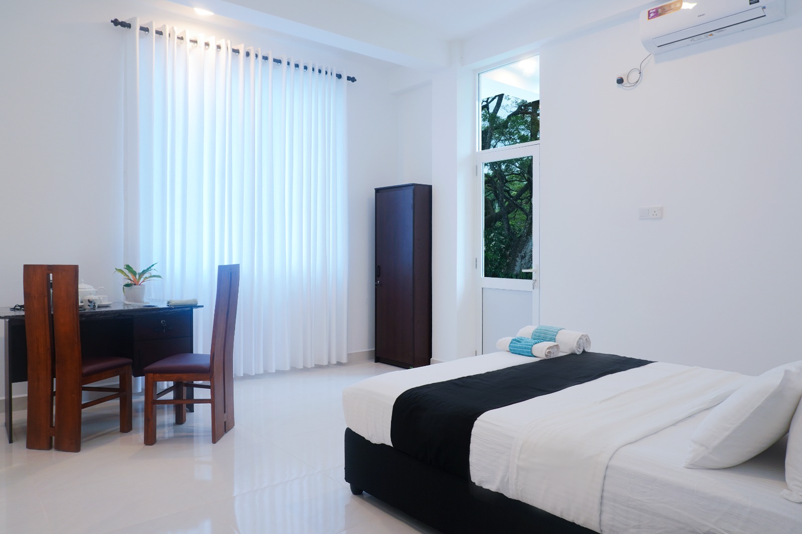 Bright guest room with a double bed, wooden writing desk and chairs, long white curtains, air conditioning and a glass door to the balcony